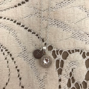 May include: Silver-toned necklace with two pendants. One pendant is a small, round charm with an engraved letter "X". The other pendant is a bullet casing with a clear crystal. The necklace hangs on a delicate chain.
