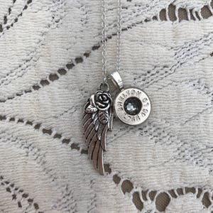 Può includere: Collana d'argento con un ciondolo che presenta una rosa e un'ala d'angelo. Il ciondolo è attaccato a un bossolo di proiettile con il testo "Shelton 9.55 45" inciso su di esso.