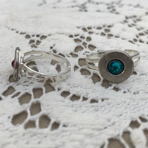 May include: Two silver-toned rings with gemstone accents. One ring features a red gemstone, while the other has a teal gemstone set within a bullet casing. The rings are displayed on a white lace surface.