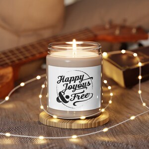 May include: A brown candle with a white label that says "Happy Joyous & Free" in a circle. The candle is lit and sitting on a wooden coaster.