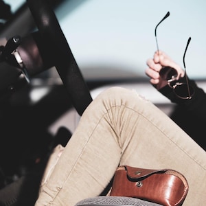 May include: A pair of dark-tinted sunglasses held in one hand, a brown leather glasses case resting on tan denim jeans, and a black steering wheel visible inside a vehicle. The scene captures a casual moment with accessories and clothing.
