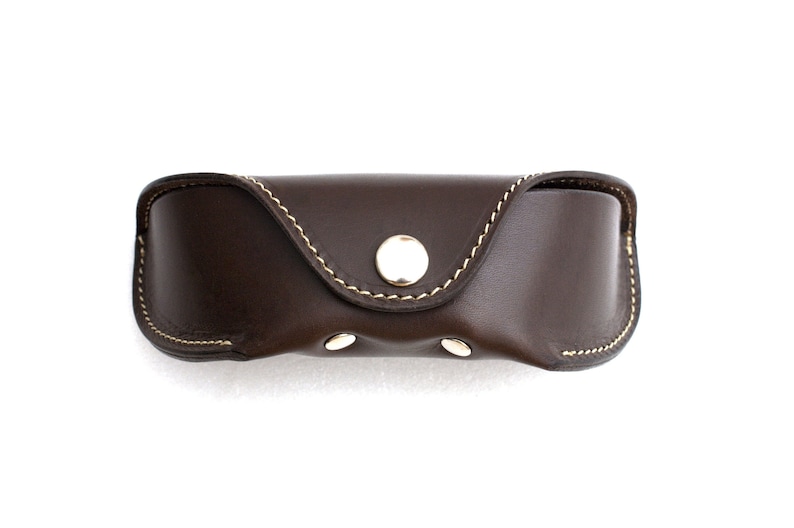 May include: A dark brown leather eyeglasses case with a curved shape and a snap closure. The case is stitched with cream-colored thread and has a gold-colored button and two smaller gold-colored rivets.