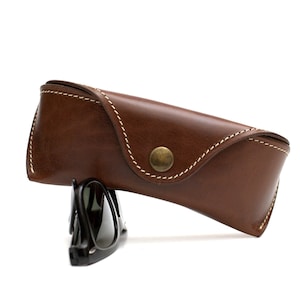Könnte beinhalten: Ein braunes Sonnenbrillenetui aus Leder mit weißen Nähten und einem Messingknopfverschluss. Schwarze Sonnenbrillen sind teilweise neben dem Etui zu sehen. Das Etui dient zum Schutz von Brillen.