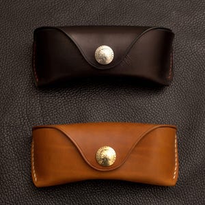May include: Two leather glasses cases, one in dark brown and one in tan, each featuring a curved flap secured by a silver-toned metal button embossed with a profile portrait and the word LIBERTY, with visible stitching along the side edges.