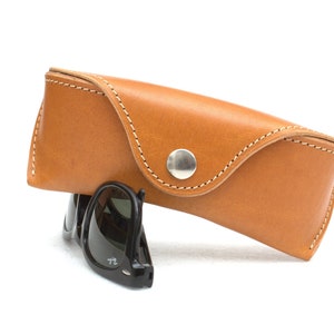 May include: A brown leather case with white stitching and a silver button closure. A pair of black sunglasses are partially visible inside the case.