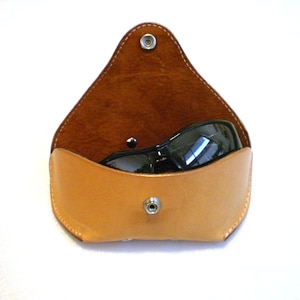 Op de afbeelding: Een bruin leren zonnebrillenetui met een drukknoop. De etui is open en er zit een zwarte zonnebril in.