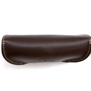 May include: A dark brown leather sunglasses case with a curved design. The case has a stitched border with gold thread and a debossed logo on the front. The case is designed to protect eyewear.
