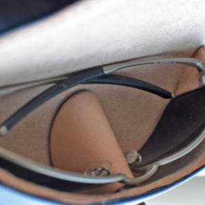 May include: Sunglasses with a silver frame and dark lenses rest inside a blue and tan leather case. The case is open, revealing the glasses and a small, tan leather pouch. The sunglasses have a sleek, modern design.
