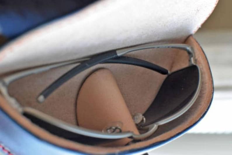 May include: Sunglasses with a silver frame and dark lenses rest inside a blue and tan leather case. The case is open, revealing the glasses and a small, tan leather pouch. The sunglasses have a sleek, modern design.