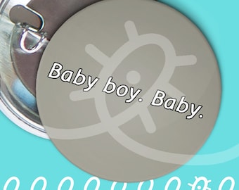 May include: A circular pin-back button with a matte gray surface featuring the white text Baby boy. Baby. A faint, light gray abstract graphic is centered behind the text. The back of the button shows a metallic pin mechanism for attachment.