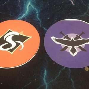 Könnte beinhalten: Zwei runde Aufkleber mit unterschiedlichen Designs. Der erste Aufkleber ist orange mit einer schwarzen Raute, die eine weiße Kobra enthält. Der zweite Aufkleber ist lila mit einer schwarzen Silhouette eines Vogels mit ausgebreiteten Flügeln und zwei gekreuzten Schwertern dahinter.