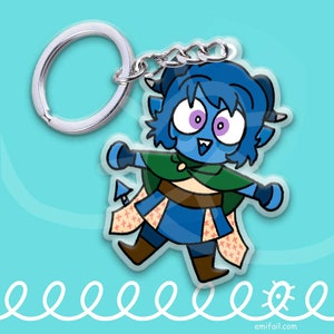 May include: A blue and green cartoon character keychain with a silver key ring. The character has large eyes, a wide smile, and a pointed tail. The character is wearing a green cloak and a brown belt.