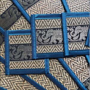Op de afbeelding: Een set geweven placemats en onderzetters met een blauwgroene rand. De placemats en onderzetters hebben een natuurlijke beige achtergrond met een zwart en zilver olifantenontwerp. De onderzetters zijn vierkant en de placemats rechthoekig.