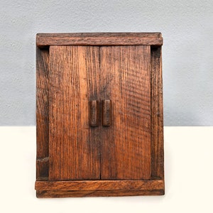 Puede incluir: Un pequeño armario de madera con dos puertas y un diseño sencillo. El armario está hecho de madera marrón oscura con un patrón de veta natural. Las puertas tienen dos pequeñas manijas rectangulares.