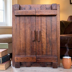 Puede incluir: Un mueble de madera artesanal con dos puertas y un diseño rústico. El mueble está hecho de madera marrón oscuro y presenta tablas verticales, una pieza superior horizontal y una base con pies. El mueble tiene dos manijas verticales.