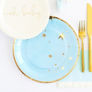 May include: A collection of baby shower party supplies. Includes a cream-colored paper plate with "oh baby" in gold script, a light blue plate with gold stars, gold cutlery, and blue and white candies.