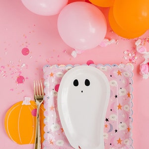 Op de afbeelding: Een roze en witte papieren bord in de vorm van een spook met een zwart gezicht en ogen. Het bord ligt op een roze tafel met oranje en roze ballonnen en sprinkels. Het bord heeft een gegolfde rand en een patroon van sterren, manen en het woord "BOO" dat wordt herhaald.