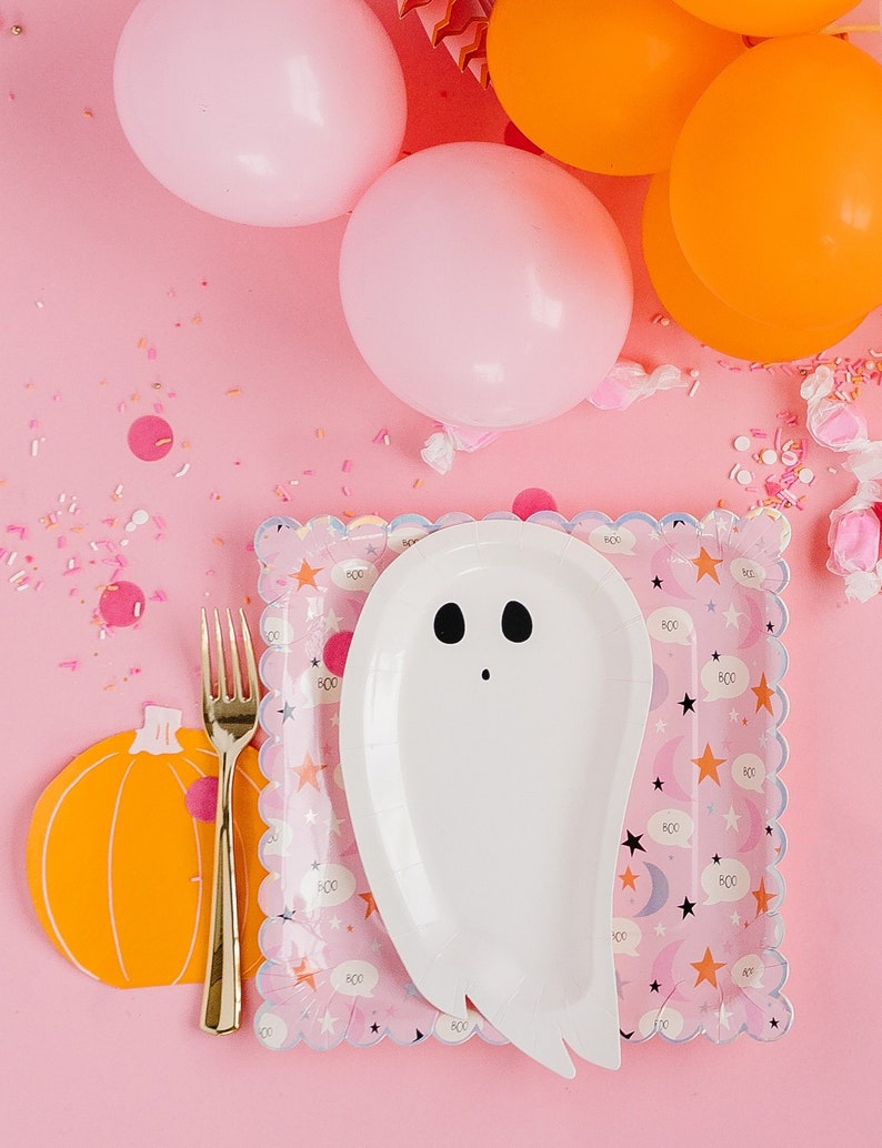 Op de afbeelding: Een roze en witte papieren bord in de vorm van een spook met een zwart gezicht en ogen. Het bord ligt op een roze tafel met oranje en roze ballonnen en sprinkels. Het bord heeft een gegolfde rand en een patroon van sterren, manen en het woord "BOO" dat wordt herhaald.