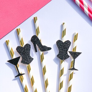 May include: An overhead shot features cocktail straws with gold stripes, and black glitter cutouts of a high heel, corset, and martini glass. Pink and white striped straws and a pink paper corner are also visible.