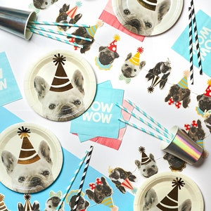May include: A party table setting with a blue and white tablecloth, white paper plates with a French bulldog wearing a party hat, and a blue and white paper banner with a repeating pattern of French bulldogs wearing party hats. The table also has blue and white striped straws, a silver cup, and blue and pink napkins with the word "WOW" printed on them.