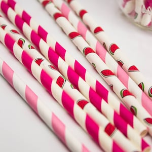 May include: A close-up of pink and white striped paper straws with a watermelon print. The straws are arranged in a fan shape on a white surface.
