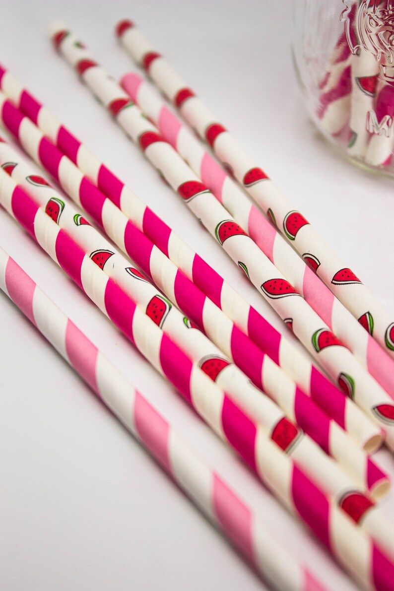 May include: A close-up of pink and white striped paper straws with a watermelon print. The straws are arranged in a fan shape on a white surface.