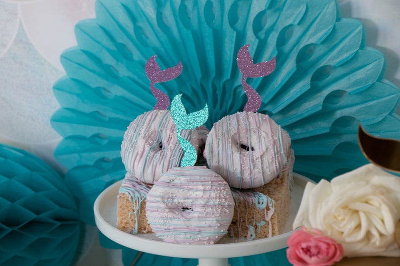 May include: A stack of four donuts decorated with white and pink frosting and sprinkles. Each donut has a mermaid tail topper with glitter accents. The donuts are on a white cake stand with a blue and white paper fan in the background.