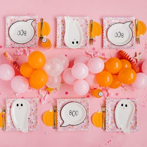 Op de afbeelding: Een roze tafelsetting met witte spook- en spraakballonplaten met het woord "BOO" erop. De tafel is versierd met oranje en roze ballonnen, confetti en kleine pompoenen.