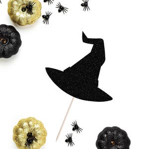 Peut inclure: Chapeau de sorcière noir pailleté avec un bâton noir, entouré de citrouilles pailletées dorées et noires et d'araignées noires sur un fond blanc.