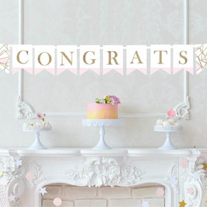Op de afbeelding: Een roze en gouden geometrische banner met het woord "CONGRATS" in gouden letters. De banner hangt boven een witte schouw met een roze en witte taart op een witte taartstandaard.