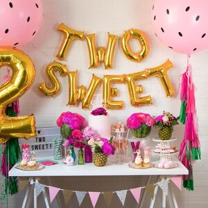 May include: A pink and gold birthday party table setting with a cake, cupcakes, and a banner that says "Two Sweet". There are watermelon and pineapple decorations, pink and green streamers, and a pink and white banner with watermelon slices.
