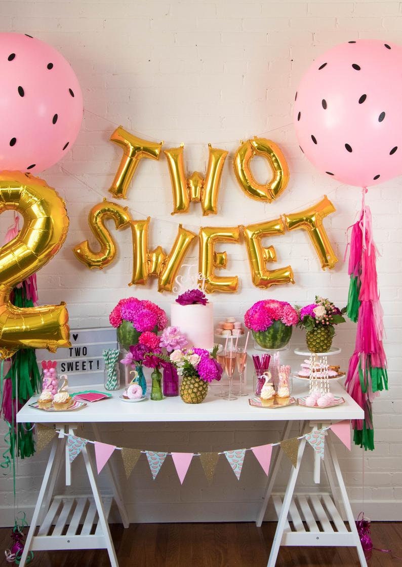 May include: A pink and gold birthday party table setting with a cake, cupcakes, and a banner that says "Two Sweet". There are watermelon and pineapple decorations, pink and green streamers, and a pink and white banner with watermelon slices.