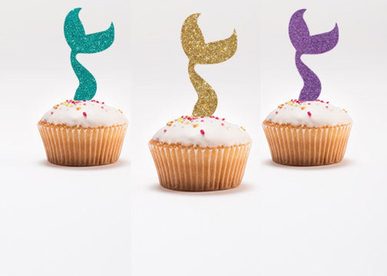May include: Three cupcakes with white frosting and colorful sprinkles topped with glitter mermaid tail cupcake toppers in teal, gold, and purple.
