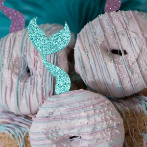 May include: A stack of pastel pink and blue frosted donuts with white sprinkles and blue glitter mermaid tail toppers.