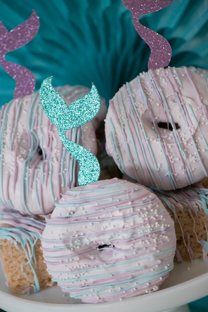 May include: A stack of pastel pink and blue frosted donuts with white sprinkles and blue glitter mermaid tail toppers.