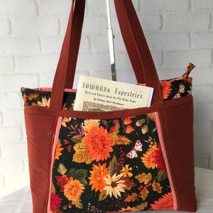 May include: A tote bag with a floral pattern featuring orange, yellow, and white flowers on a black background. The bag has rust-colored sides and straps, with a pink accent. A book titled "Towanda Tapestries" is visible inside the bag.