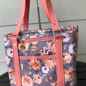 May include: A floral tote bag with a gray background and peach-colored flowers. The bag has peach-colored straps and accents, a zippered top, and a small charm. The bag is designed for carrying items.