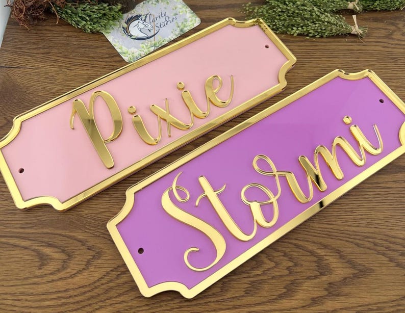 May include: Two rectangular nameplates with gold trim and lettering. One is pink with the name "Pixie" in gold script, and the other is purple with "Stormi" in gold script. The nameplates are on a wooden surface.