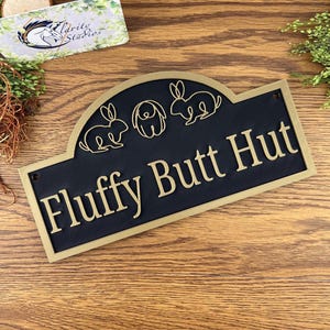 May include: Black and gold metal sign with a rounded top and the text "Fluffy Butt Hut". The sign features three small animal silhouettes: a dog and two rabbits.