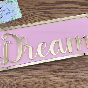 May include: A pink sign with a gold border and the word "Dream" written in gold cursive lettering.