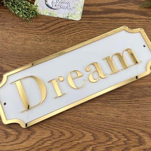 May include: A rectangular white sign with gold trim and the word "Dream" in gold letters. The sign has a decorative shape and is mounted on a wooden surface. The sign is a decorative item.