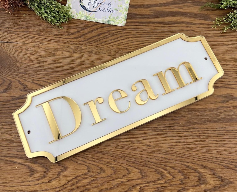 May include: A rectangular white sign with gold trim and the word "Dream" in gold letters. The sign has a decorative shape and is mounted on a wooden surface. The sign is a decorative item.