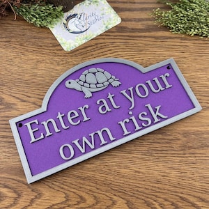 May include: A purple sign with a silver border and the text "Enter at your own risk" in silver lettering. A silver turtle is at the top. The sign is rectangular with an arched top.