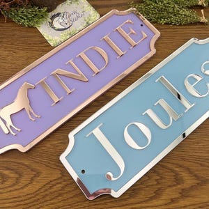May include: Two rectangular nameplates. One is lavender with rose gold trim and lettering, and a horse silhouette. The other is light blue with silver trim and lettering. Both have rounded corners.