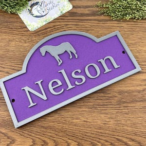 May include: A purple sign with the name "Nelson" in raised silver lettering, and a horse silhouette. The sign has a grey border and two small holes for hanging. The sign is likely for a stable.