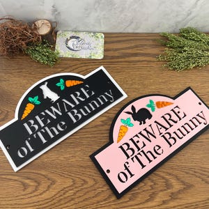 May include: Two decorative signs with the text "BEWARE of The Bunny". One sign is black with a white bunny and carrots, the other is pink with a black bunny and carrots. The signs are on a wooden surface.