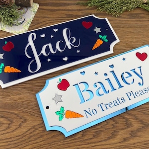 May include: Two decorative nameplates in navy blue and white. One says "Jack" with carrot, apple, and star designs. The other says "Bailey No Treats Please!" with similar designs. Both have a scalloped edge.