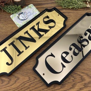 May include: Two rectangular signs with black borders and gold and silver mirrored surfaces. The word "JINKS" is displayed on the gold sign, and "Ceasar" is on the silver sign. The signs are on a wooden surface.
