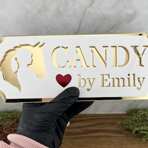May include: White sign with gold trim and a gold horse silhouette. The sign reads "CANDY by Emily" with a red heart.