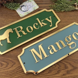 May include: Two rectangular horse nameplates. One is green with "Rocky" in gold lettering and a gold horse silhouette. The other is blue-green with "Mango" in gold lettering. Both have gold trim.
